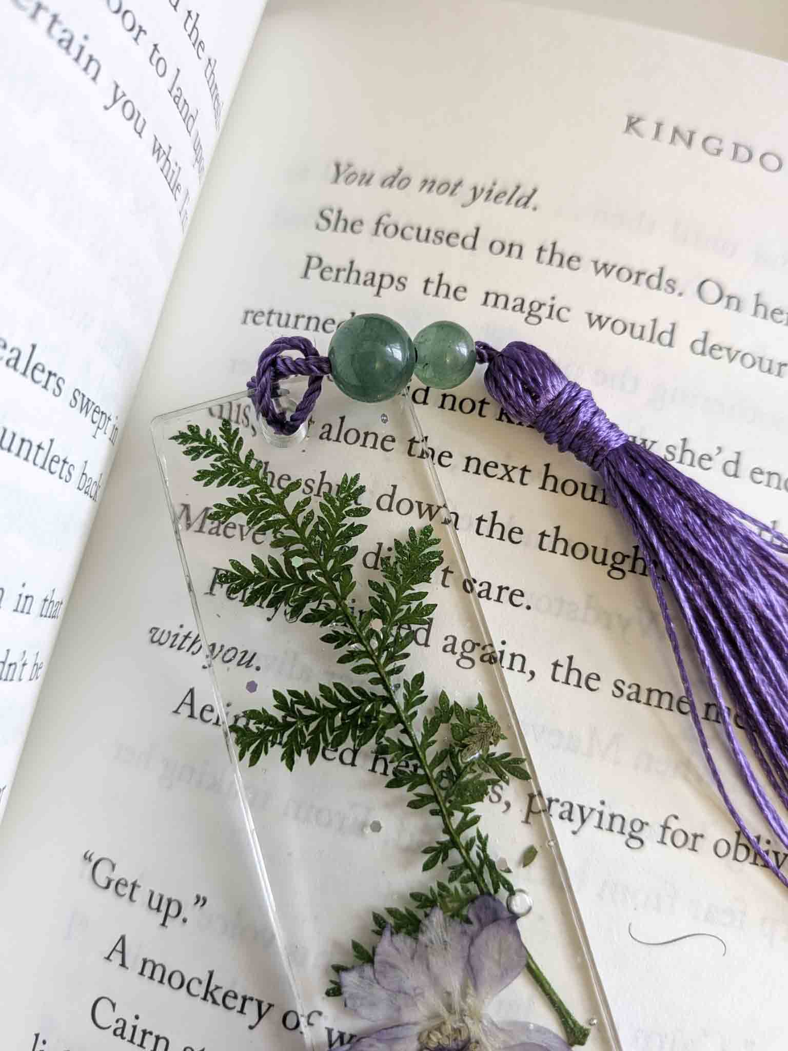Decorative bookmark with green leaves, purple flowers, and a tassel on an open book.